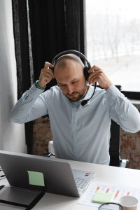 Bald man in blue shirt putting on headset at desk, working remotely on laptop in a modern call center.